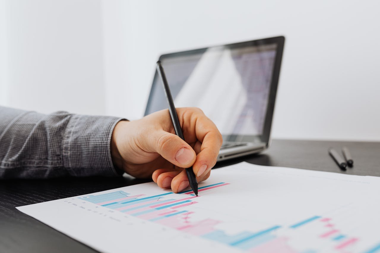 Hand holding a pen analyzing a performance chart on paper, with a laptop in the background, illustrating Conversion Rate Optimization strategies for improving website performance and user engagement.