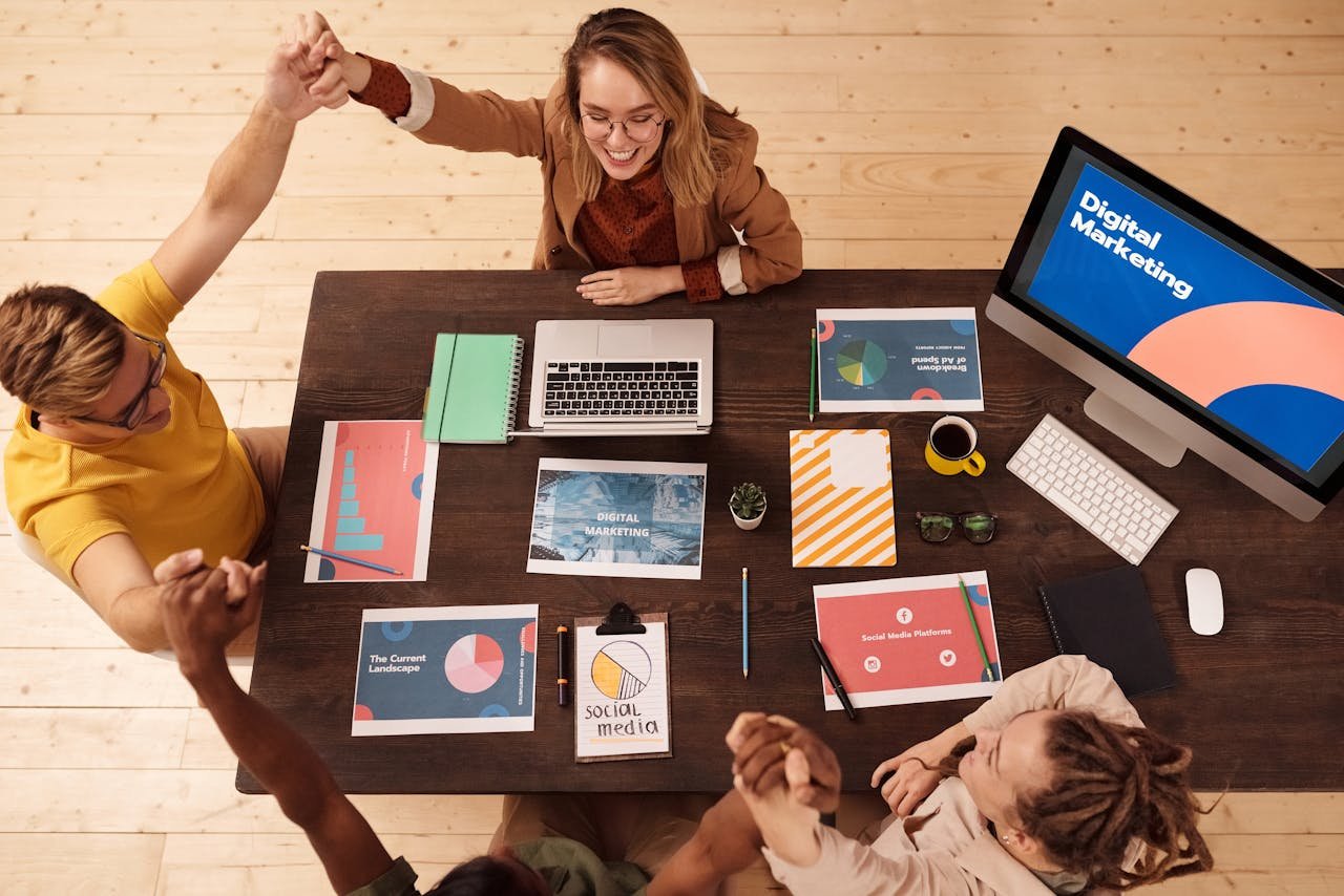 Team celebrating success in a collaborative workspace, surrounded by digital marketing materials, including charts and reports, with a computer displaying "Digital Marketing."