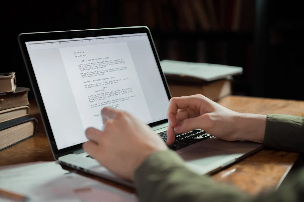 Person typing on a laptop with a visible script document, surrounded by books and papers, illustrating content creation and strategic messaging for effective audience engagement.